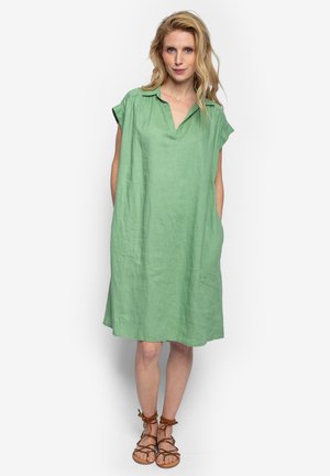 Blonde Frau steht und trägt ein lockeres, grünes knieumspielendes Kleid mit kurzen Ärmeln und braue Schnürsandalen vor einem weißen Hintergrund.