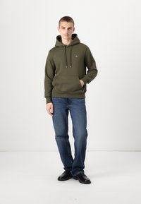 Olivengrønn hettegenser med justerbare snorer, frontlomme og ribbet mansjetter, kombinert med blå jeans og svarte sko. Enkel design.