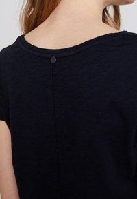 Námořnická modrá blůza s krátkým rukávem, kulatým výstřihem a zapínáním na knoflík na zádech. Jemná textura s lehkým leskem a švy.