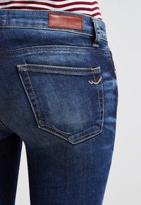 Jeans de mezclilla azul con una vista detallada de los bolsillos traseros, que presentan una etiqueta de cuero y un ligero desgaste. Costura durable y tela texturizada.