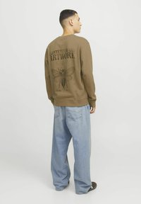 Sudadera marrón con un gran gráfico negro de una polilla y las palabras "ARTWORK CONTEMPORÁNEO" en la parte de atrás. Pantalones de denim azul de corte holgado.