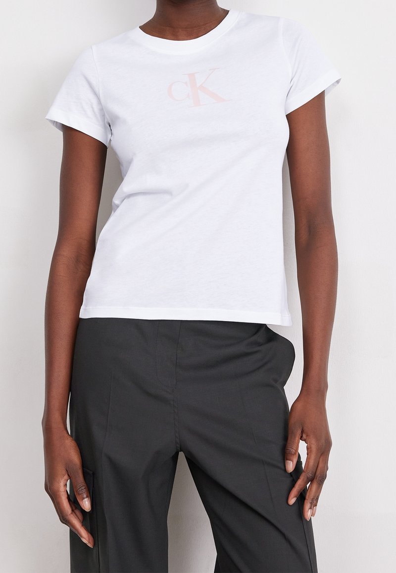 Persona vestida con una camiseta blanca de manga corta con un sutil logo "cK" y pantalones negros de cintura alta, de pie frente a un fondo blanco.