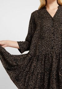 Czarna sukienka z rękawami o długości trzech czwarte, w kropkowany wzór w odcieniu beżowym, z zapięciem na guziki z przodu oraz rozkloszowanym dołem z falbaną. Miękki, lekki materiał.