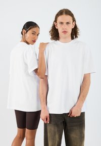 Két ember visel fehér, oversized pamut pólót; az egyik elölről, a másik hátulról látható. Az ingek laza ujjaikkal és kerek kivágásukkal tűnnek ki.