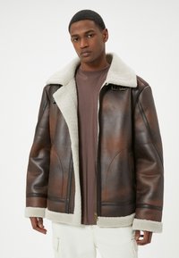 Giubbotto in pelle marrone con colletto in pile beige, chiusura a zip e tasche frontali. Presenta una finitura texturizzata e dettagli di cucitura visibili.