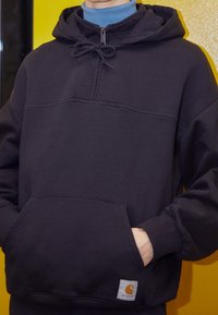 Felpa con cappuccio di un blu scuro navy, con una tasca frontale, design a mezza zip e cordino regolabile. Presenta il logo Carhartt in fondo.