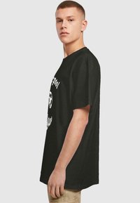 Zwarte katoenen t-shirt met korte mouwen, voorzien van witte tekst en een voetbalafbeelding op de voorkant. Relaxte pasvorm, iets oversized ontwerp.