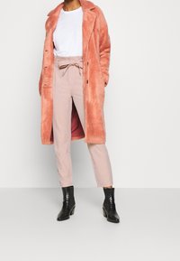 Rosa fuskpälsjacka över vit t-shirt, parat med högmidjade beige byxor och svarta klackade ankelskors. Slät textur, avslappnad design.