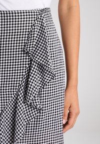 Schwarz-weißer Vichykaro-Rock mit Rüschendetail, ausgestattet mit einer figurbetonten Taille und leichtem Material. Eine Hand ist seitlich sichtbar.