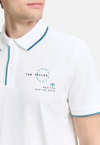 Hvid polo skjorte med en turkise og sorte stribede krave. Har et trykt rundt design med tekst "TOM TAILOR" og "REG TRM SEAS AND SKIES."