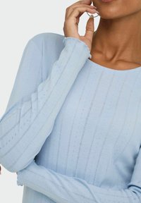 Eine Nahaufnahme einer Person in einem hellblauen, gerippten Langarmshirt, die ihr Kinn auf ihre Hand stützt und eine entspannte Pose einnimmt.
