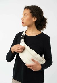 Baumwoll-Schultertasche in Creme mit einem Reißverschluss, verstellbarem Tragegurt und geprägt Logo. Das Modell hält die Tasche vor einem schwarzen Shirt.