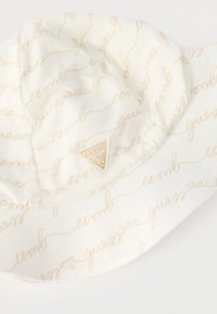 Λευκό καπέλο τύπου κουβά με μπεζ καλλιγραφικό τύπωμα «Guess 1981 USA» και μικρό τρίγωνο λογότυπο Guess στο μπροστινό μέρος.