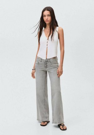 Vit ribbad linne med framknappar, kombinerad med ljusgrå vida jeans och svarta flip-flops, stående mot en neutral bakgrund.