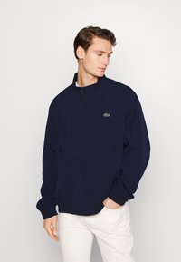 Mornarško modra zip-up jakna z visokim ovratnikom in diskretnimi elastičnimi manšetami, ki ima majhen zeleni logotip na prsih in stranske žepke.