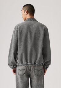 Γκρι τζιν μπουφάν με ελαστικό τελείωμα και μανσέτες, με γιακάκο λαιμόκοψη και χαλαρή γραμμή. Η πίσω όψη δείχνει ελάχιστες λεπτομέρειες και ραφές.