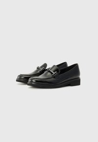 Mocassins en cuir noir avec une finition brillante, dotés d'une sangle simple et d'un accent métallique. La semelle plate légèrement surélevée offre de la stabilité.