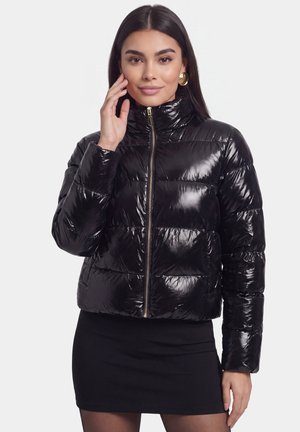 Giovane donna con lunghi capelli scuri, che indossa orecchini d’oro, una giacca imbottita nera lucida e una gonna nera, posando con la mano vicino al volto.