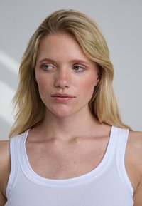 Blonde vrouw met een lichte huid en blauwe ogen, gekleed in een wit hemdje, kijkt naar links tegen een effen lichtgrijze achtergrond.