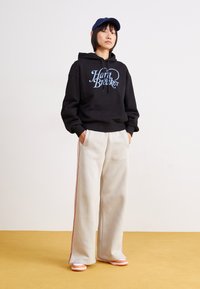 Svart hettegenser med teksten "Haarit Braker", beige vide bukser med røde sidestriper og lysfargede sneakers. En uformell sportswear look.