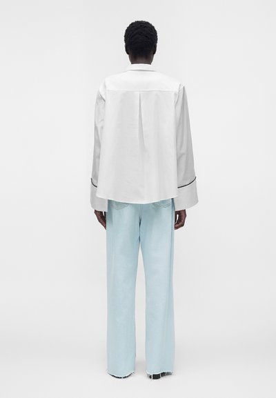 Personne de dos portant une chemise blanche ample à manches longues avec un liseré noir et un jean large bleu clair avec des ourlets effilochés.