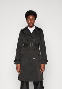 Dunkelgrüner Trenchcoat mit doppelreihiger Knopfleiste, großen Knöpfen, tailliertem Gürtel und breiten Revers. Mit Schulterklappen und Taschen.