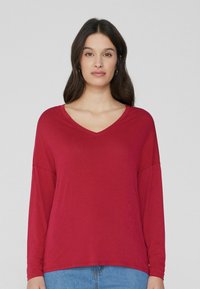 Rotes Langarmshirt mit V-Ausschnitt, aus weichem Stoff gefertigt. Lockerer Schnitt mit abgesenkten Schultern, kombiniert mit blauen Jeans.