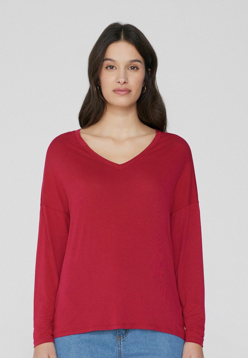 Rotes Langarmshirt mit V-Ausschnitt, aus weichem Stoff gefertigt. Lockerer Schnitt mit abgesenkten Schultern, kombiniert mit blauen Jeans.