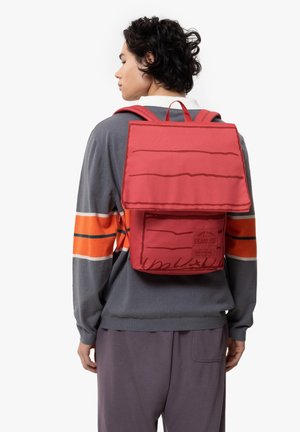Mochila de lona roja con una solapa estructurada, que presenta una superficie texturizada y detalles gráficos impresos. Correas ajustables y un asa superior.