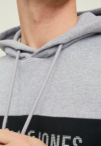 Šedá mikina s kapucňou, vybavená šnúrkou, textúrovanou látkou a kontrastným čiernym pásom s bielymi pruhovanými nápismi na spodnej strane.