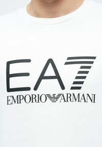 Λευκό βαμβακερό t-shirt με μεγάλο μαύρο σχέδιο που απεικονίζει το "EA7" πάνω από το "EMPORIO ARMANI", διακοσμημένο με ένα στυλιζαρισμένο λογότυπο αετού.