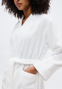 Γυναίκα που φοράει λευκό ρόμπα από πετσέτα με ζώνη, υφασμένα μανίκια και κεντημένο λογότυπο "RL" στο στήθος, με το χέρι στην αριστερή τσέπη.