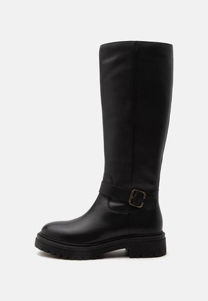 Bota negra de cuero hasta la rodilla con suela gruesa y una correa con hebilla metálica alrededor del tobillo, mostrada de lado.