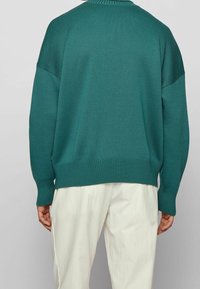 Un pull en tricot turquoise avec des épaulettes tombantes et un bord côtelé, associé à un pantalon ample de couleur claire. La texture est tissée et lisse.