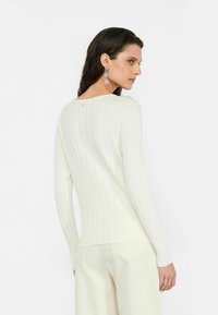 Top bianco a maniche lunghe con costine e una leggera lucentezza, scollatura rotonda e un piccolo logo vicino alla schiena. Indossato con pantaloni chiari.