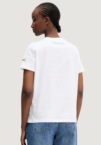 Camiseta de algodón blanca con un corte relajado, que presenta mangas cortas y un pequeño detalle bordado en negro en la manga izquierda.