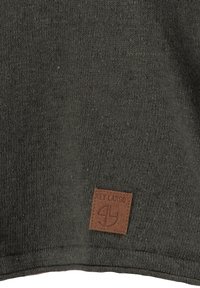 Dunkelgrüner Strickstoff mit strukturierter Oberfläche. Ein braunes Lederetikett mit "Key Largo" und Logo befindet sich in der unteren Ecke.
