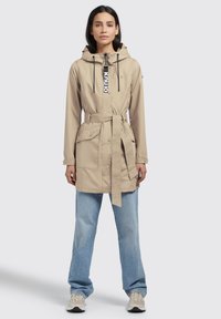 Beige lightweight jacket with a hood, drawstrings, belted waist, and front pockets, paired with light blue jeans and sneakers.