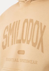 Sudadera con capucha beige con texto desvanecido "SMILodox", "2013©" y "ESSENTIAL SPORTSWEAR" impreso en el frente.