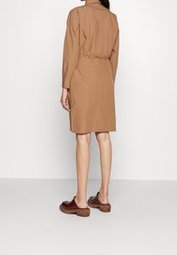 Abito camicia marrone realizzato in un tessuto leggero, con colletto, maniche lunghe e cintura in vita, abbinato a zoccoli marroni.