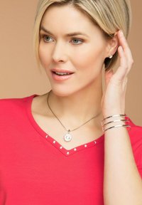 T-shirt rossa a collo a V con borchie argentate lungo il collo, abbinata a un bracciale argento e a una collana con pendente ottagonale. Tessuto dalla superficie liscia.