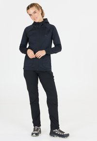 Svart atletisk jacka med huva, dragkedja fram och sidofickor, som är kombinerad med svarta byxor och trailskor med grå accenter och snören.