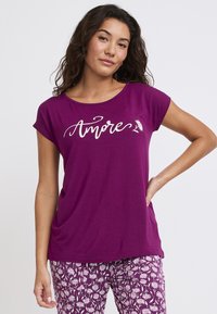 Fioletowa koszulka z krótkim rękawem o luźnym kroju, z napisem "Amore" w białym stylu. Połączona z wzorzystymi fioletowymi spodniami.