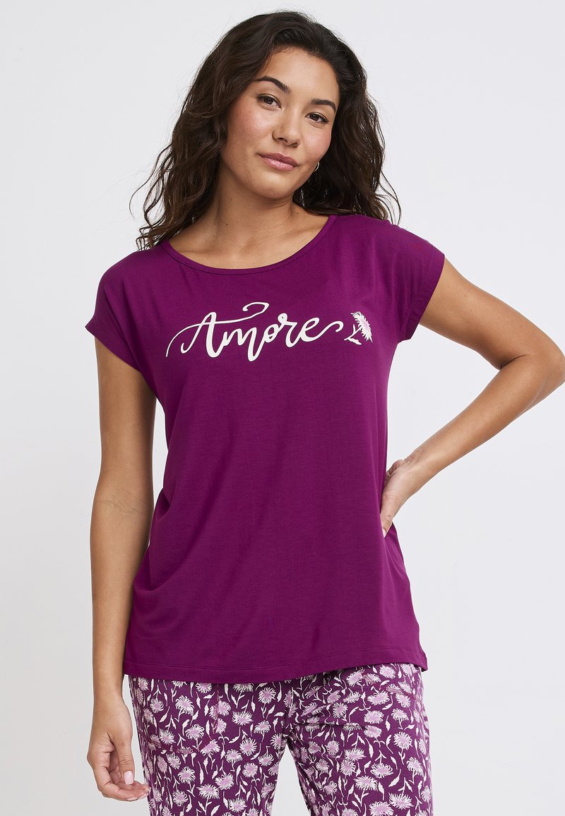 Fioletowa koszulka z krótkim rękawem o luźnym kroju, z napisem "Amore" w białym stylu. Połączona z wzorzystymi fioletowymi spodniami.