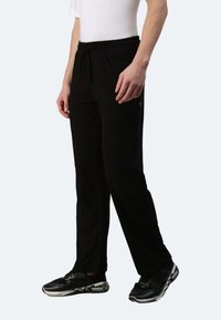 Schwarze Sporthose mit einem Kordelzugbund, Seitentaschen und einem glatten, leichten Material. Kombiniert mit schwarzen Sneakers.