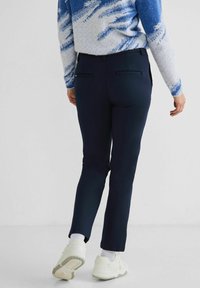 Pantaloni tapered blu navy con una texture liscia, dotati di due tasche posteriori. Indossati con un maglione a righe blu chiaro e bianco e sneakers bianche.