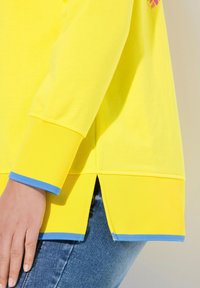 Haut en coton jaune vif avec un ourlet incurvé, des fentes sur les côtés et des accents bleus aux poignets. Les détails de couture visibles mettent en valeur le design.
