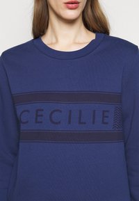 Felpa blu navy con scollo arrotondato, design a righe testurizzate e la parola "CECILIE" in lettere scure sul petto.