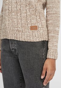 Beige gebreide trui met een kabelpatroon, voorzien van een leren label. Gepaard met donkergrijze jeans. Brei textuur zichtbaar op de trui.