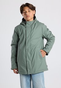 Green waterproof jacket with a hood, snap closures, and side pockets. Smooth texture and relaxed fit. Worn over light blue jeans.